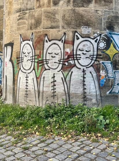 Graffiti von Maurice Sendaks „Wilden Kerlen“ auf dem Sandsteinpfeiler einer Brücke. Davor Pflastersteine und Gras. Das Graffiti ist noch gut erhalten, daneben sind weitere Sachen zu sehen. Die drei Figuren sind weiß und schwarz gearbeitet.