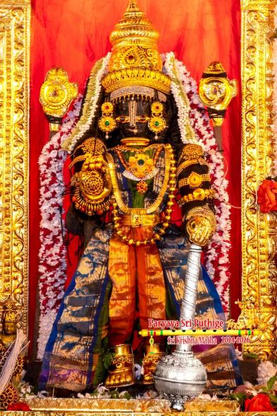 Close up of an idol of Hindu deity Krishna in red pants, blue pant flares, green-and-black op; standing with right hand in blessing pose, left hand holding the top of a mace's handle (the mace head rests on the floor); wearing gold shoes, gold palm coverings, gold crown, and lots of gold ornaments. A string of white and red flowers (mixed in a dot pattern) around Krishna floor-floor. A gold icon of "chakra" (disc) behind Krishna's right; an icon of "shankha" (conch) behind on the left. The sides of a gold arch behind Krishna is visible (but not the entire arch). Assorted loose flowers at Krishna's feet.
