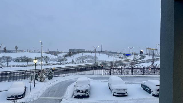 Verschneiter Parkplatz, teilweise geräumt. Die Autos sind von einer dicken Schneeschicht bedeckt.
Dahinter verschneite Hügel und Strassen.