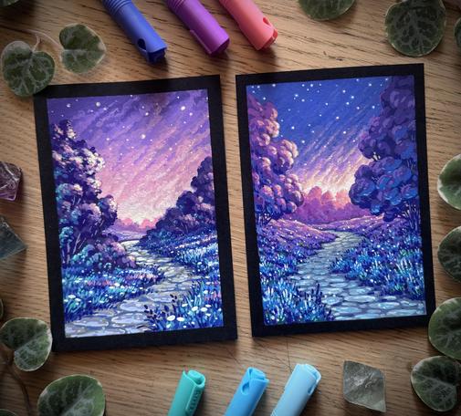 A photo of two DinA6 acrylic marker paintings on black paper, placed side by side on a wooden desk and surrounded by acrylic markers, fluorite crystals, and leaves. Both sceneries feature a winding path through wooded grassland in pink/purple/blue tones with a dreamy, otherworldly sunset vibe. The starry sky on the left is more purple, while the one on the right features some blue as well. The grass texture on the left is more „classic“, while the one on the right looks rather like lavender.