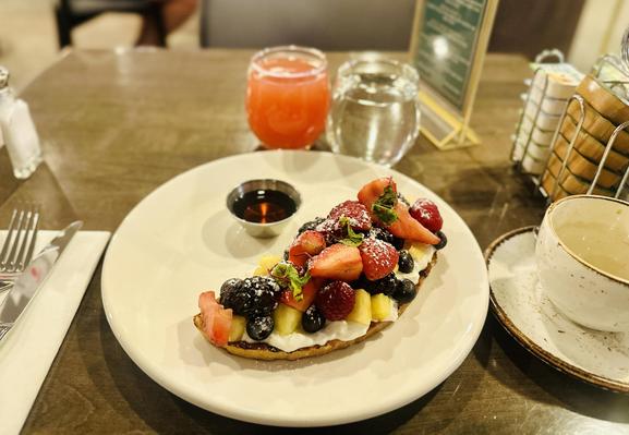 Ein Geller mit einem halbmondförmigen Brot, das mit vielen Erdbeeren, Brombeeren und Himbeeren, sowie weiteren Früchten belegt ist. Daneben ein Schälchen Ahornsirup. Dazu Wasser, Grapefruit-Saft und eine leere Tasse Kaffee.