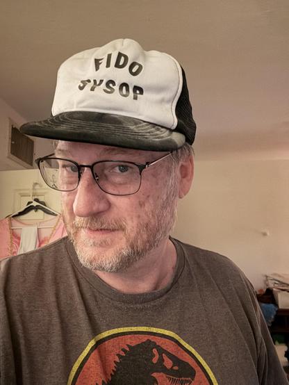 A person is wearing a white and black trucker hat with the text “FIDO SYSOP” on it. They are also wearing a brown T-shirt featuring the Jurassic Park logo with a dinosaur silhouette. The background appears to be a casually furnished room with clothes hanging and shelves visible.

FidoNet is a worldwide computer network designed for communication between bulletin board systems (BBSes), reaching its peak popularity in the 1990s. It operated using a store-and-forward system, which allowed email and public forum messages (as well as files) to be exchanged between participating BBSes, many of which only supported one user at a time. Messages were transferred in bundles between systems—usually at night to minimize long-distance phone charges—using published routing lists called nodelists. FidoNet was platform-independent, eventually comprising tens of thousands of nodes globally, organized into zones and nets to efficiently route mail and manage network traffic. This system was a vital part of early online communities before the Internet became widely accessible.