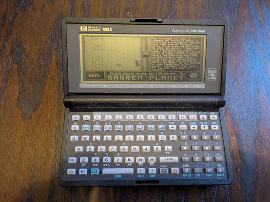 A photograph of a Hewlett Packard 100LX running the default Barren Planet campaign First Landing, showing the Nuvutech briefing for the first mission.