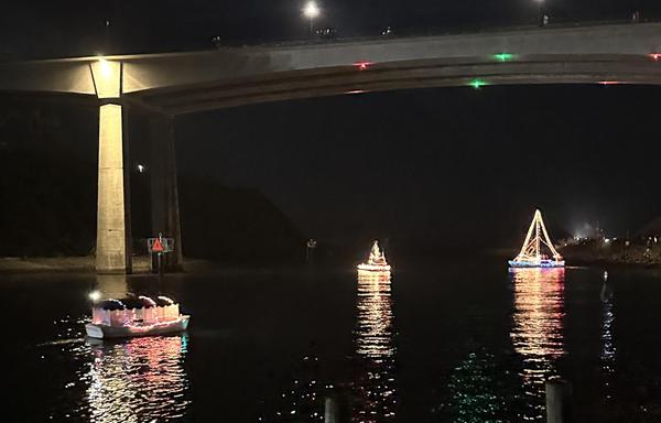 Boats decorated with colorful holiday lights float under a bridge at night during a festive parade, their reflections glowing on the dark water.
