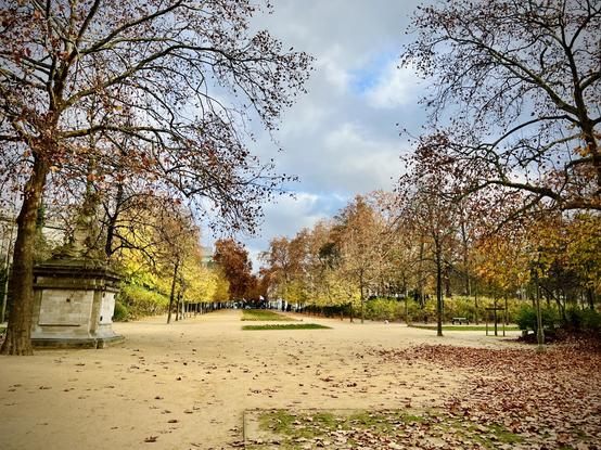 This image depicts a wide, open park area, resembling a large square or clearing. The ground is a mix of dirt and short grass, liberally sprinkled with fallen brown and yellow leaves. Trees with mostly bare branches and some patches of pale yellow leaves frame the perimeter of the open space. On the left side, a stone monument or pedestal with ornate carvings stands under one of the trees. In the background, beyond the open area, more trees and hints of buildings are visible under an overcast sky. The overall impression is one of a spacious, quiet park in late autumn or early winter.