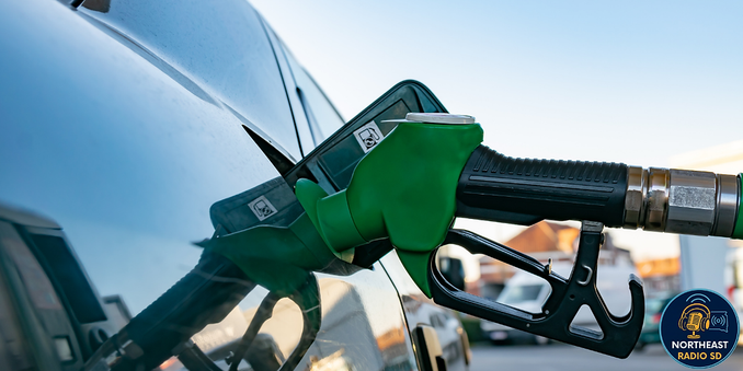 Green fuel nozzle in a silver car's gas tank, outdoors on a sunny day. Logo with "NORTHEAST RADIO SD" in the bottom right corner.