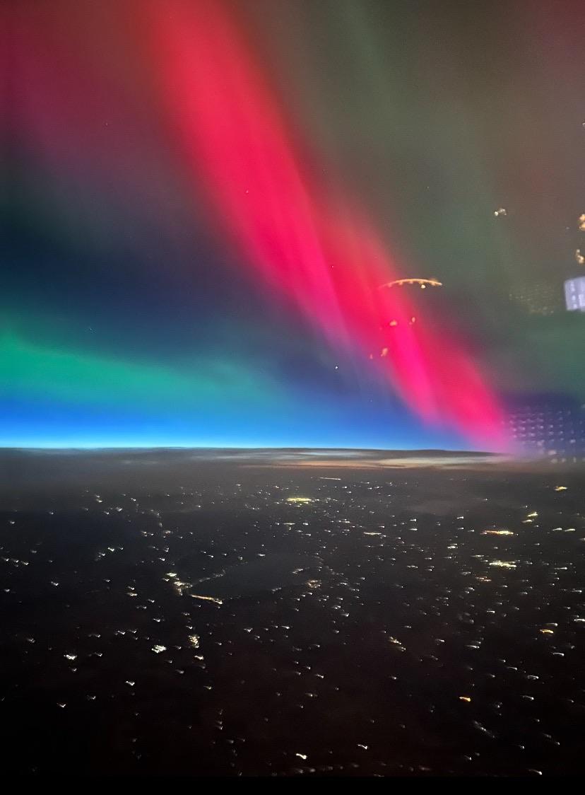 The image shows a vibrant display of the aurora borealis, featuring vivid red and green lights against a dark sky. Below, a landscape is visible with scattered city lights, suggesting it is an aerial view taken from an aircraft.