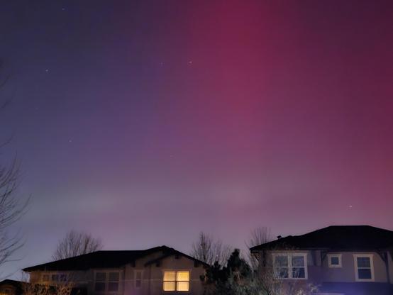 The night sky over houses. There is a pink hue in the top right part of the picture with some light greenish horizontal streaks near the bottom.