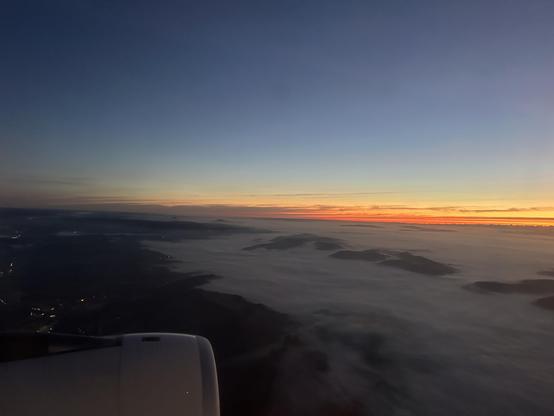 Blick aus dem Flugzeugfenster. Morgenrot über dem Westen. Am Boden dicker Nebel rund um die Hügel. In der linken Ecke sieht man das linke Triebwerk.
