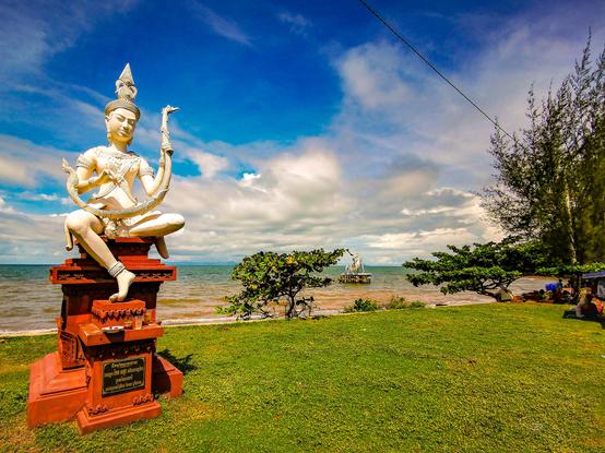 Photo shows a white statue of an Apsara lady demigod playing a Pin, an arched harp instrument. She is placed on a red fundament standing on a strip of green turf. In the background there is a a beach and sea stretching out. Out at sea another statue of a blue swimming crab is visible on a plattform. It is a sunny day, and most of the sky is deep blue. But in the horizon there are some light clouds showing.  Cambodia is as safe, comfortable and kind as ever. Angkor, Kep, Kampot warmly welcomes visitors.