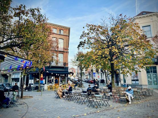 This photo captures a bustling city square or plaza with numerous people seated at outdoor tables and chairs, suggesting a cafe or restaurant area. The ground is paved with cobblestones and scattered with fallen autumn leaves. On the left, a building with a dark awning houses a cafe, with its outdoor seating area partially covered by a glass or transparent canopy adorned with string lights. Several people are sitting at tables in this area. To the right and extending into the center, many empty black metal chairs and small tables are arranged on the cobblestones, waiting for patrons. Large trees with golden-yellow autumn leaves are prominent on both sides of the square. In the background, multi-story brick buildings line the street, and more people can be seen walking or gathering further down the street. The sky is overcast, and the overall atmosphere is that of an active urban space in autumn.