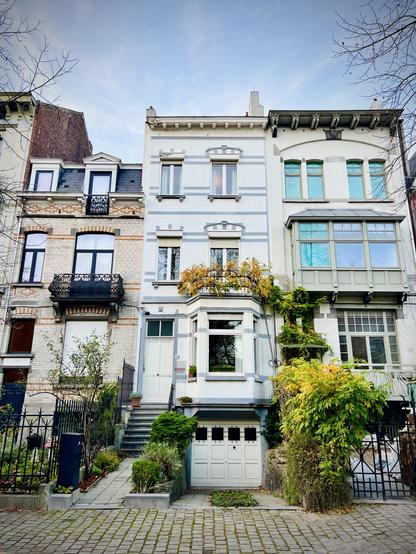 This image displays a row of three distinct townhouses, characteristic of European urban architecture, under a muted, overcast sky. The central house is light grey with white trim, featuring bay windows on the ground and first floors, and a small balcony on the first floor adorned with some climbing, yellow-leaved plants. A set of concrete steps leads up to its main entrance. Below the steps is a white garage door. To its left, a building with light brickwork and black wrought-iron balconies stands. To the right, another building with a light-colored facade features a large bay window on the upper floor and a climbing green plant covering part of its front. All three houses have multiple windows and intricate details, and a black metal fence with decorative elements runs along the front of the properties, bordering a cobblestone sidewalk.