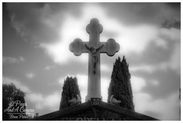 A monochrome (black and white) photograph by Bron Peirce captures a detailed, stone crucifix with the figure of Christ (corpus) on a tombstone.
The cross has trefoil like ends and is silhouetted against a bright, cloudy sky, which is a significant part of the composition.
Two dark, columnar cypress trees stand behind the tombstone, flanking the base of the cross. Two small stone bird figures (likely doves) are visible on the top corners of the tombstone base beneath the cross.