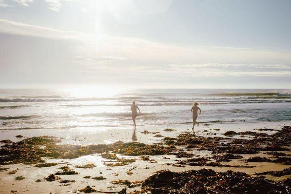 Two people run into the sea at a beach during the North East Skinny Dip event, silhouetted against the bright sunlight.