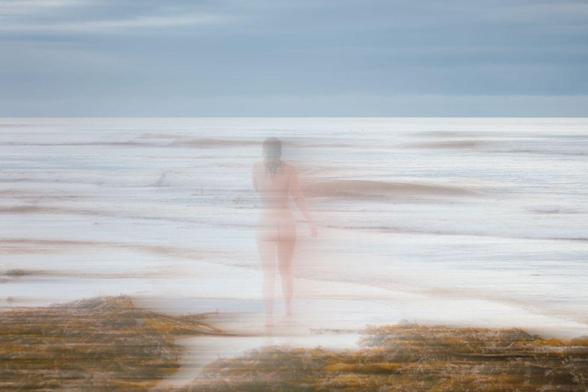 A person walks into the sea, viewed from behind, with seaweed in the foreground and a cloudy sky above.