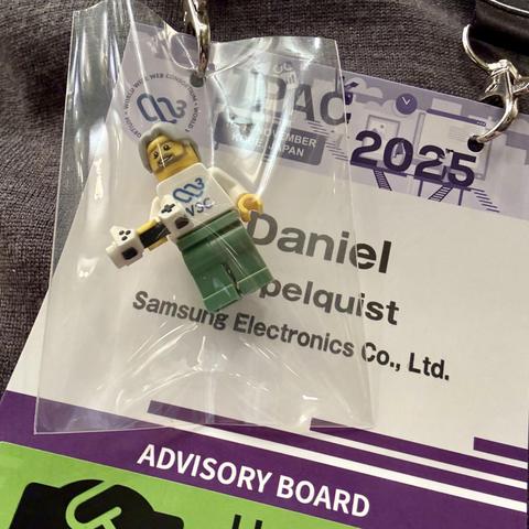 A LEGO figure in a clear plastic bag, holding a video game controller, with a w3c logo sticker on it, is attached to a name badge. The badge displays the name "Daniel" and mentions "Samsung Electronics" along with the event details for "TPAC 2025”.