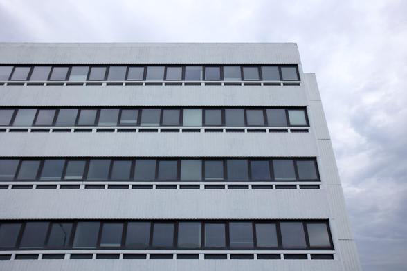 A modern white building with flat, horizontal rows of rectangular, dark-framed windows. The facade is simple and geometric, stretching across several floors. The sky above is overcast, with grey clouds dominating the background.