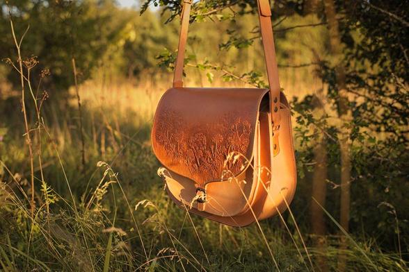 Light colored bag hangs on a tree branch by it's shoulder strap. Sunlight falls onto the bag from the right side and brings out the nice deepness of the carved decorations on the bag's cover flap. This bag has a large main compartment and additional smaller pocket right under the flap in front of the main compartment. Photo is taken on the meadow near the forest at summertime.