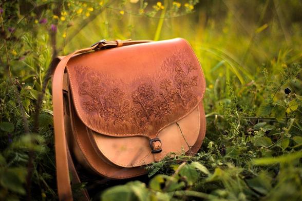 Light colored bag is set onto the ground. There are meadow-flowers and some dewberries amongst other plants. This photo presents the front of the bag. There is a smaller buckle for a fastening and the shoulder strap also has a buckle. The edges of the bag are stitched by light coloured thread.