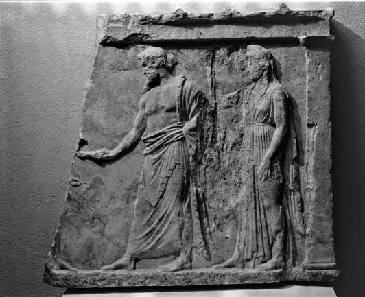 Marble votive relief to Zeus and Hera. The relief is framed on the right by a pillar with base and capital supporting an entablature crowned by a flat roof composed of tiles. The left side of the relief has been broken off. A bearded Zeus wears a himation wrapped to leave his chest bare. He has a long pointed beard and his long hair is gathered up into a bun at the back of his head. Zeus faces left and extends a phiale (shallow bowl) towards another figure, now missing. The only evidence of thi…