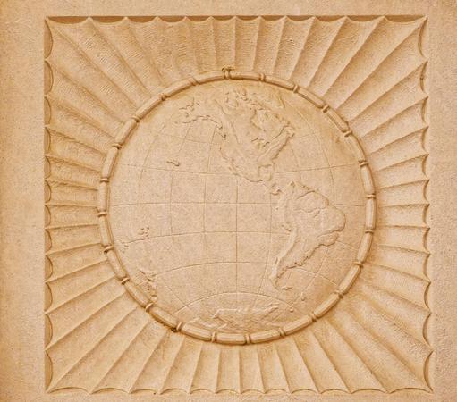 On a light yellowed white background rock is carved an image of the globe centering north America surrounded by what looks like a circle of long beats and outside that a set of scalloped ridges inscribed in a square