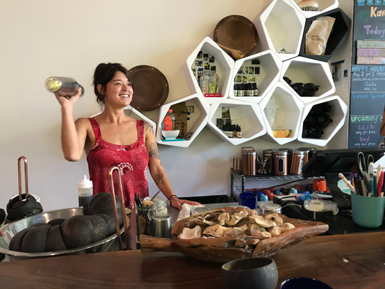 A young woman stands behind a counter with a wooden top. She is smiling, and shaking a cocktail shaker in her right hand. Her black hair is worn up, and she's wearing a red, satiny camisole. On the bar is a bowl of coconut shells stacked (for drinking kava) and a bowl of dry kava root, used to explain where kava comes from.