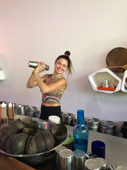 A young woman behind the bar at Melo Melo kava bar. She is smiling as she shakes a cocktail shaker with two hands. She's wearing black leggings and a tightly fitting, sleeveless top. On the bar is a bowl of coconut shells, and various shakers and mixing implements.