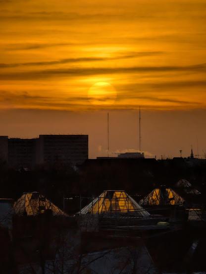 A dramatic, deep orange sunrise over a city skyline. The sun is a large, hazy orb centered in the frame, illuminating high-level clouds. In the darker foreground, the silhouettes of urban buildings and several glass pyramid structures reflect the intense glow.