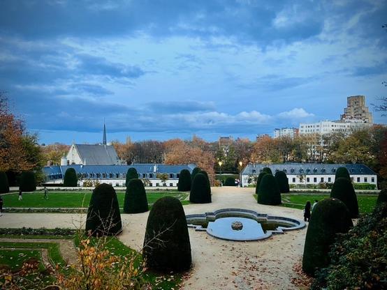 This image captures a wide view of a formal European garden on an overcast day.
In the foreground, there are tan gravel paths covered with a scattering of fallen brown and yellow leaves. On either side of the paths, and further into the midground, are numerous tall, dark green, conical, and bell-shaped evergreen hedges, meticulously pruned. In the center of the midground, a multi-tiered circular fountain with still water reflects the cloudy sky. Several people are visible walking along the gravel paths and on the grass.
Beyond the immediate garden features, the midground opens up to expansive, well-maintained green lawns. Behind these lawns, a long, low, white building with a gray, pitched roof and numerous dormer windows stretches across the frame. To the left of this building, a taller, lighter-colored building with a prominent steeple suggests a church or chapel.
The background is filled with a mixture of trees, some still green and others showing autumn colors of yellow and brown. Above the trees and buildings, a wide, cloudy sky dominates the upper portion of the image, giving the entire scene a soft, diffused light. To the far right, a distinctive, light-colored, modern, stepped building stands out against the horizon, contrasting with the more traditional architecture of the other buildings. The overall impression is one of peaceful, well-maintained grounds under a muted sky.