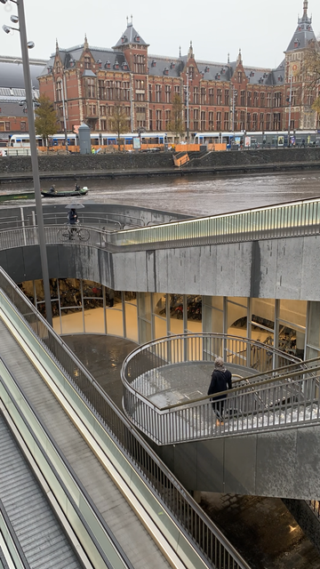 View of stairs and escalators descending several floors below water level (a small boat is visible). In the background, white and blue trams in front of a large station building.