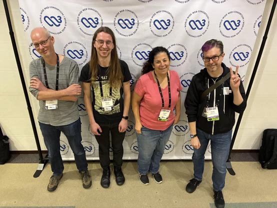 A group of four individuals stands in front of a backdrop featuring the World Wide Web Consortium (W3C) logo. They are smiling and posing together. The setting appears to be a conference or event, with name badges visible on their attire.