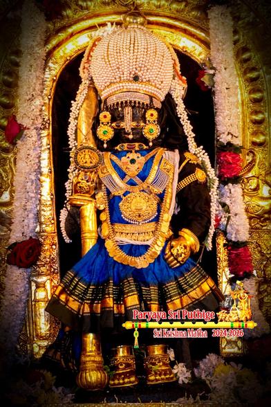 Closeup of idol of Hindu deity Krishna in blue (black and gold lined border) standing, a gold-covered staff in right hand, left hand on front left waist; wearing gold shoes, gold palm coverings, a dome-shaped pearl crown, and lots of gold ornaments including a long necklace made from gold coins. An ornate gold arch with broad sides behind Krishna, but the arch is not fully visible. A string of white flowers (some red roses) hang down from somewhere above Krishna and flow to floor on both sides of Krishna. A string of jasmine buds (that look like pearls) come out from behind the pearl crown and flow down on both sides of Krishna to his waist.