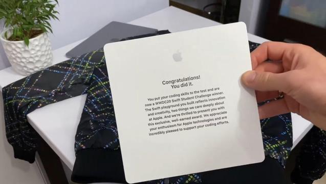Hand holds a square Apple congratulations card above a patterned jacket on a desk, with a potted plant and laptop in the background.