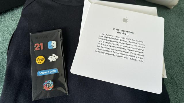 Set of WWDC pins on black fabric beside an Apple congratulations card celebrating a Swift Student Challenge/WWDC21 award.