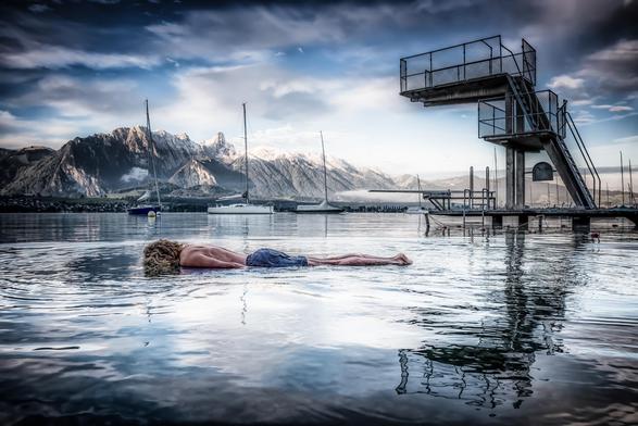 Ein Mensch liegt bäuchlings auf einer spiegelglatten Wasserfläche, als würde er auf dem Wasser schweben. Er trägt eine blaue Badehose, der Oberkörper ist nackt, die Haare sind lang und blond. Im Hintergrund ragen schneebedeckte Berge in den Himmel, davor liegen einige Segelboote ruhig auf dem See. Rechts steht ein hoher Sprungturm mit mehreren Plattformen und Geländern, der sich im Wasser spiegelt. Das Licht ist weich, die Wolken am Himmel leicht dramatisch gefärbt, wodurch eine ruhige, fast surreale Stimmung entsteht.