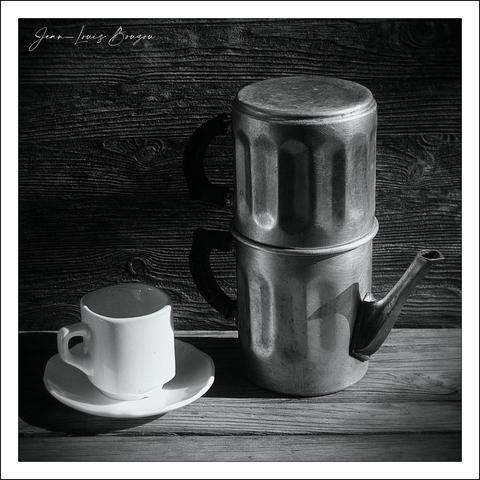 On the left edge of the image, a small white coffee mug made of ceramic sits on a matching white saucer. The mug exhibits a single handle on the left side and is positioned upright on the saucer. On the right side of the image, a very old, old-fashioned, double-walled metal coffee maker sits on the surface, similar to the one depicted in the classic French film Faustina. Although the maker is not noted on the scene, it resembles the style of one made by Maison Polar Café.
The objects are illuminated by strong light originating from the right-hand side of the makeup room. The left places directly in the shade, and the objects create awesome shadows on the wooden surface. The left section captures shadows on the side so that the wooden surface appears to have two distinct parts. This results in disagreement between the shadows and the shadows on the left, providing a stark contrast to the light and shadows on the right.
The background showcases a planked wood surface made of two crossed strips, with tightly spaced vertical planks in a light finish of pine, fittingly called redwood planks. Another it seems to be lurking behind it. Aside from the background being much darker and showing a disruption over some of the distance of the surface.