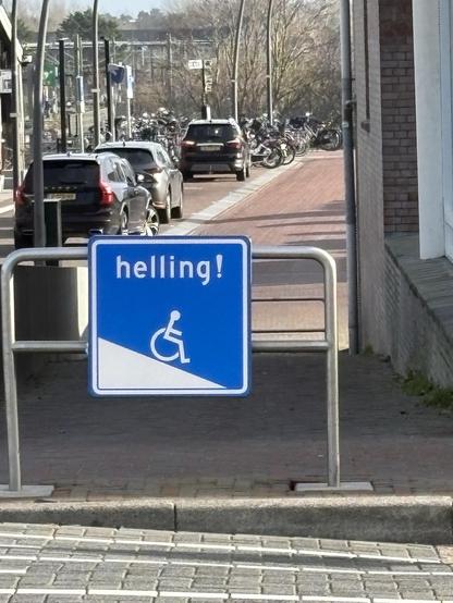 Blauw verkeersbord met een een rolstoel op een helling en de tekst ‘helling!’ voor een stuk aflopende straat.