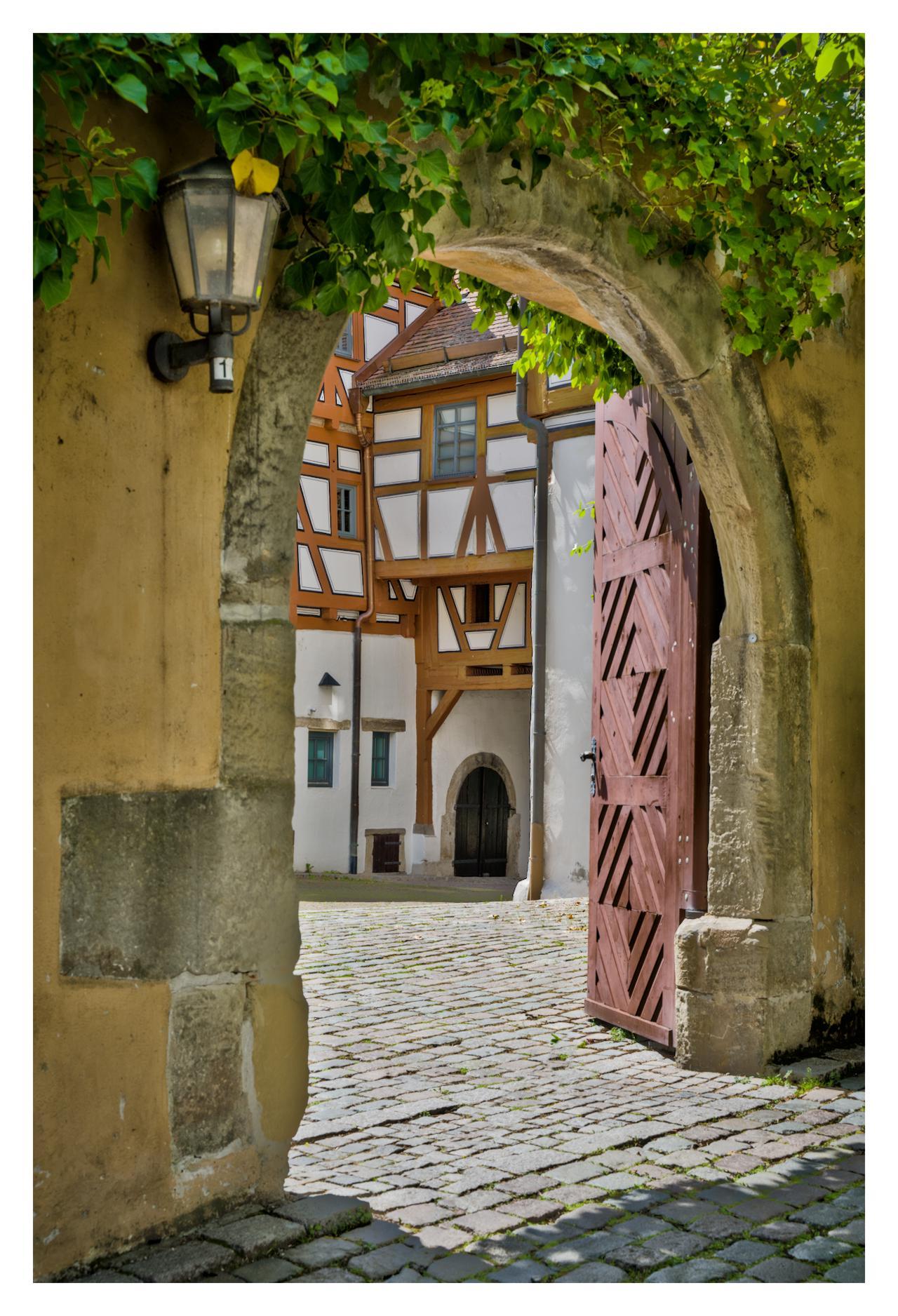 A stone archway frames the scene, covered in lush green ivy vines that drape over its curved top. On the left side of the arch hangs a vintage black lantern with a yellow bulb, marked with the number "1." Through the arch, a cobblestone pathway leads into a sunny courtyard. Ahead stands a traditional half-timbered building in German style, with white plaster walls crisscrossed by dark wooden beams forming geometric patterns. A large, open wooden gate with a zigzag design in reddish-brown slats reveals a small arched doorway beyond. The ground is paved with uneven gray cobblestones, dotted with patches of grass, and sunlight casts soft shadows, creating a quaint, historic atmosphere.