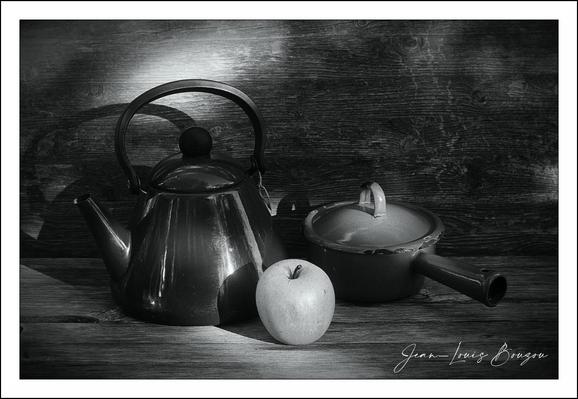 This black-and-white image beautifully captures a quiet still life scene set against a textured wooden backdrop. The composition includes a polished metal kettle, a classic cooking pot with a lid, and a smooth apple positioned in the foreground.

The kettle, with its gently curved spout and robust handle, reflects subtle highlights that emphasize its shiny surface, suggesting it's made of stainless steel or similar material. Next to it, the cooking pot, which appears to be cast iron or enameled, features a side handle and a round lid with a wide knob, evoking a sense of homemade comfort and domesticity.

The apple, nestled between these two sturdy kitchen items, contrasts with their metallic nature through its soft, organic shape. Its smooth skin and light tone provide a focal point of calm and natural beauty among the more industrial objects.

The monochrome palette adds a contemplative, timeless quality to the image, stripping away distractions and focusing attention on form, light, and shadow. This style often evokes themes of simplicity, stillness, and the passage of time—reminding viewers of the quiet rituals embedded in everyday life.
