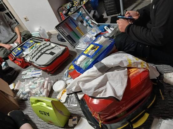 2 Notfallrücksäcke liegen auf dem Boden sowie ein offener Notfallkoffer, ein AED (Defibrillator) steht auch daneben, viele Verbandsmaterialien überall im Bild verteilt, man sieht 3 Leute (die beine) auf dem Boden sitzen, im Schneidersitz und diese Checken das Material.