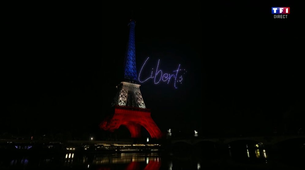 A night view of the Eiffel Tower illuminated in the vertical blue, white, and red colors of the French flag. To the right of the tower's upper section, the word "Liberté" (Liberty) is spelled out in white cursive lights against the black sky. The red lights at the base of the tower are reflected in the dark water of the river below