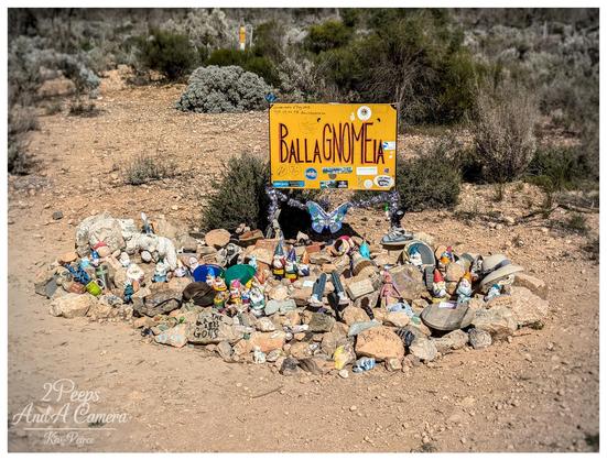 A wide angle photo of a yellow wooden sign in a dry, scrubby roadside environment. The sign, hanging between two posts wrapped in fairy lights, reads "BALLAGNOMEIA" in large orange, whimsical text.
Below the sign is a large cairn or pile of rocks covered in dozens of small garden gnomes and various trinkets, including a blue butterfly decoration, a small toy goat, and some hats. The ground is dry dirt and gravel.