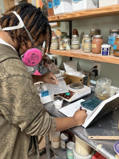 A person looking down, writing in a note book, wearing a dust mask. They are surrounded by glaze ingredients and bottles of terra sig, a scale and a cup filled with white powder (glaze ingredients.) 