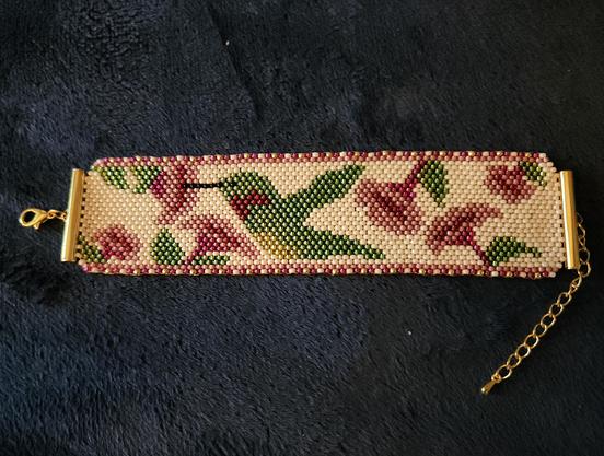 A brick stitch beadwork bracelet with a cream base. In the center of the design is a ruby throated hummingbird surrounded by pink and red trumpet flowers.