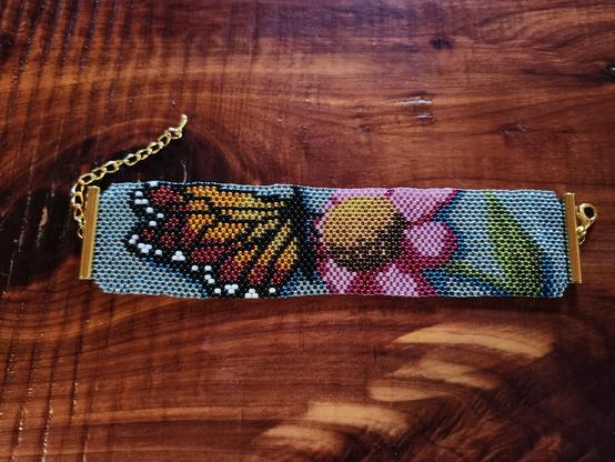 A brick stitch beadwork bracelet in a light blue background if a monarch butterfly perched on a pink coneflower.