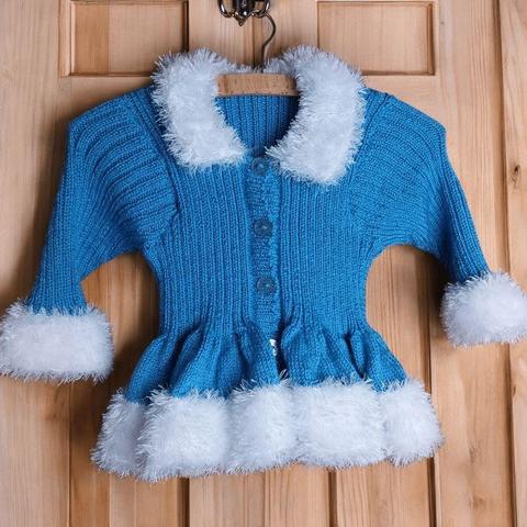 A bright blue knitted baby cardigan with fluffy white trim on the collar, cuffs, and hem hangs on a wooden hanger against a light wood panel door background.

