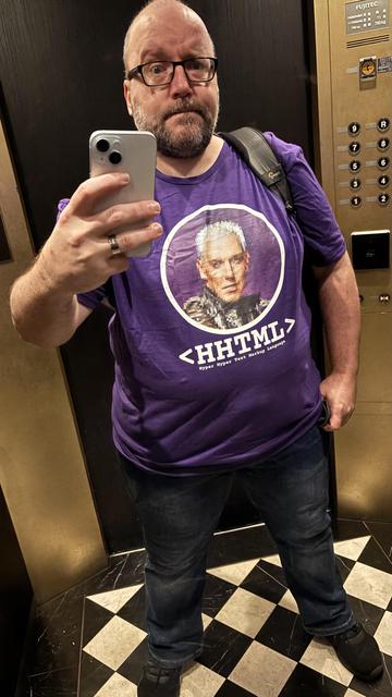 selfie in hotel elevator, wearing a purple tshirt with a large pixelated face - the lead guy from band Scooter - in a circle; underneath, in chunky white letters:

<HHTML>
Hyper Hyper Text Markup Language
