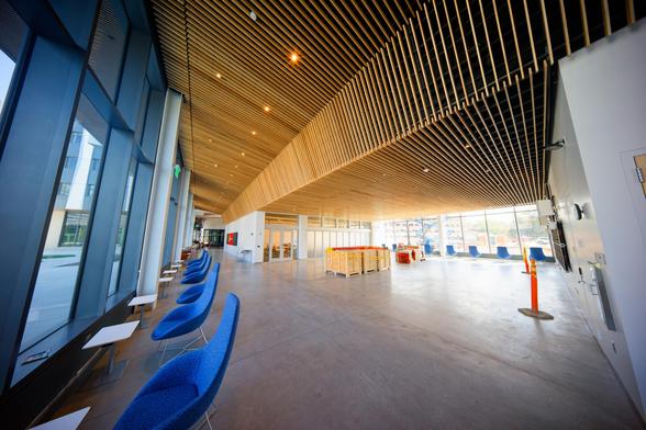 A row of blue chairs faces towards the window at the far left,  the ceiling is made of wood slats and slants down across the center to flat ceiling hall to the right