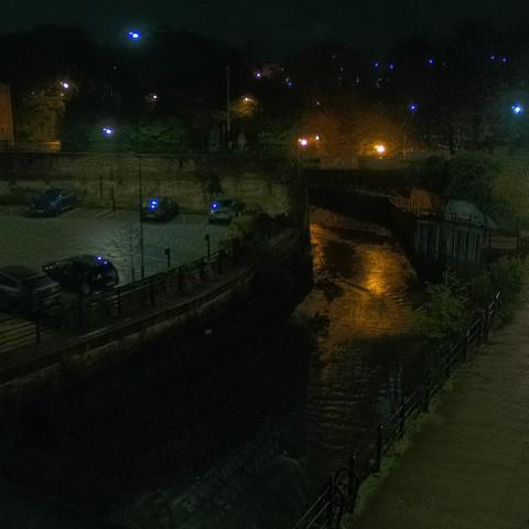 Photograph of the tidal stretch of Ouseburn River at south side of Ouseburn Bridge in Newcastle upon Tyne. To the left (south-west) of the bridge is Cut Bank, and to the right (north-east) is Byker Bank. In the foreground is the pedestrian Riverside Walk and the photo also spans some surrounding built-up areas and trees, with sky in the background. Still image captured from webcam at 03:12 hrs on Friday 14 November 2025.