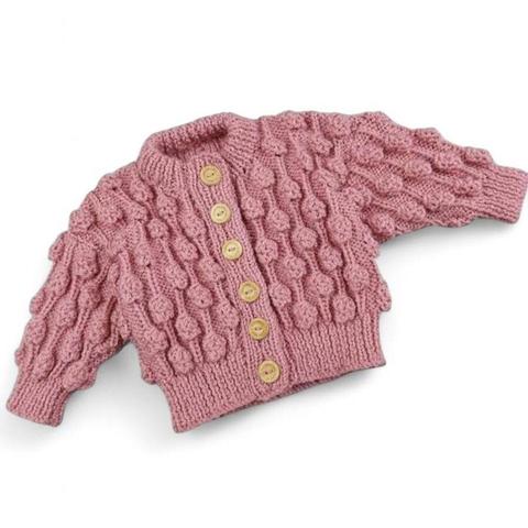 The image shows a hand-knitted, pink baby cardigan with a textured bubble pattern and wooden buttons down the front. It has ribbed cuffs, hem, and collar. The background is plain white.